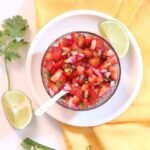 A bowl of strawberry jalapeno salsa on a white plate on a yellow tablecloth with garnish on the side